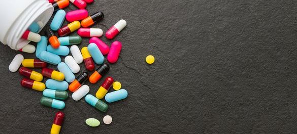 Image features a collection of multicoloured pills spilling out from container, on black counter. 